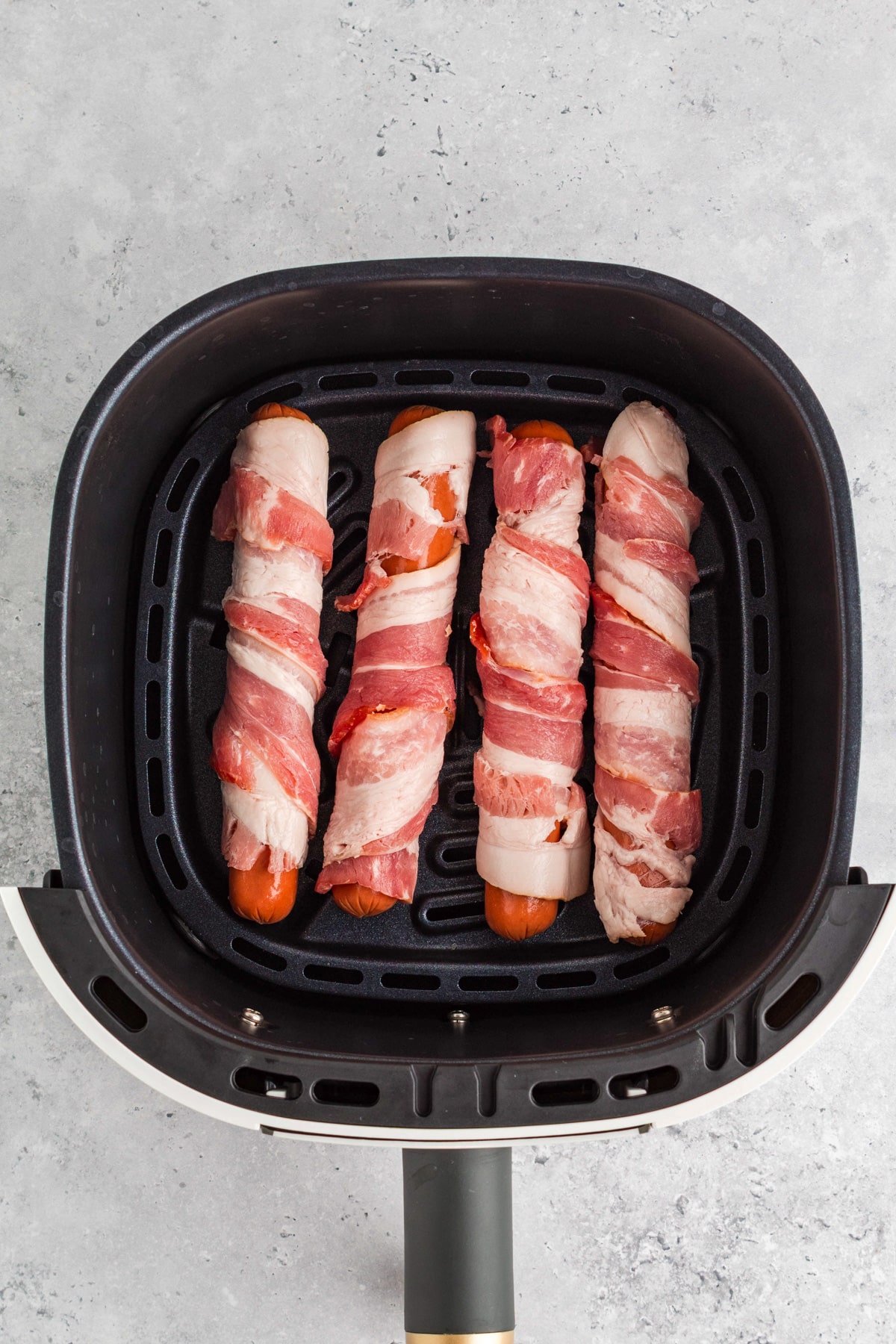 Four hot dogs wrapped in bacon are placed evenly in an air fryer basket, ready to be cooked.