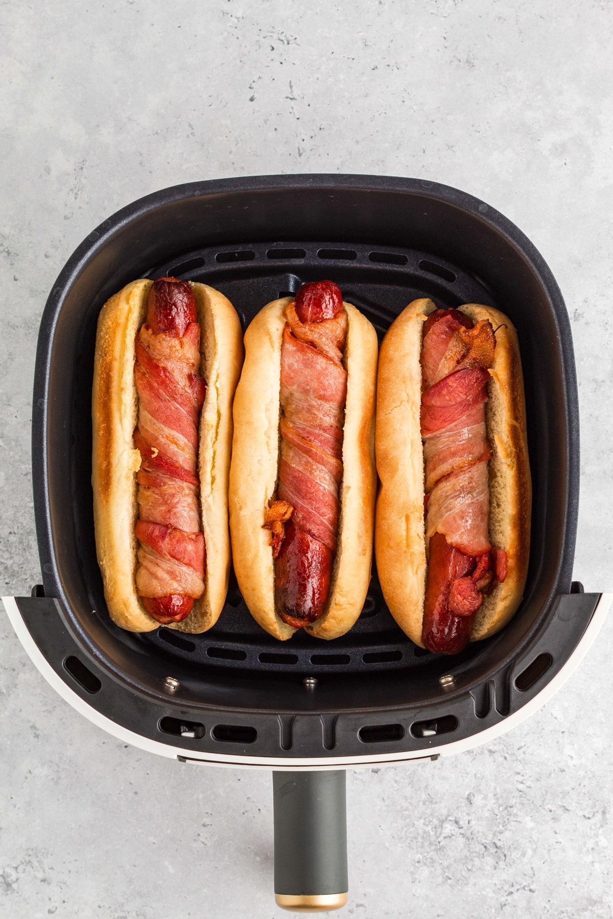 Three bacon-wrapped hot dogs in buns side by side inside an air fryer basket.