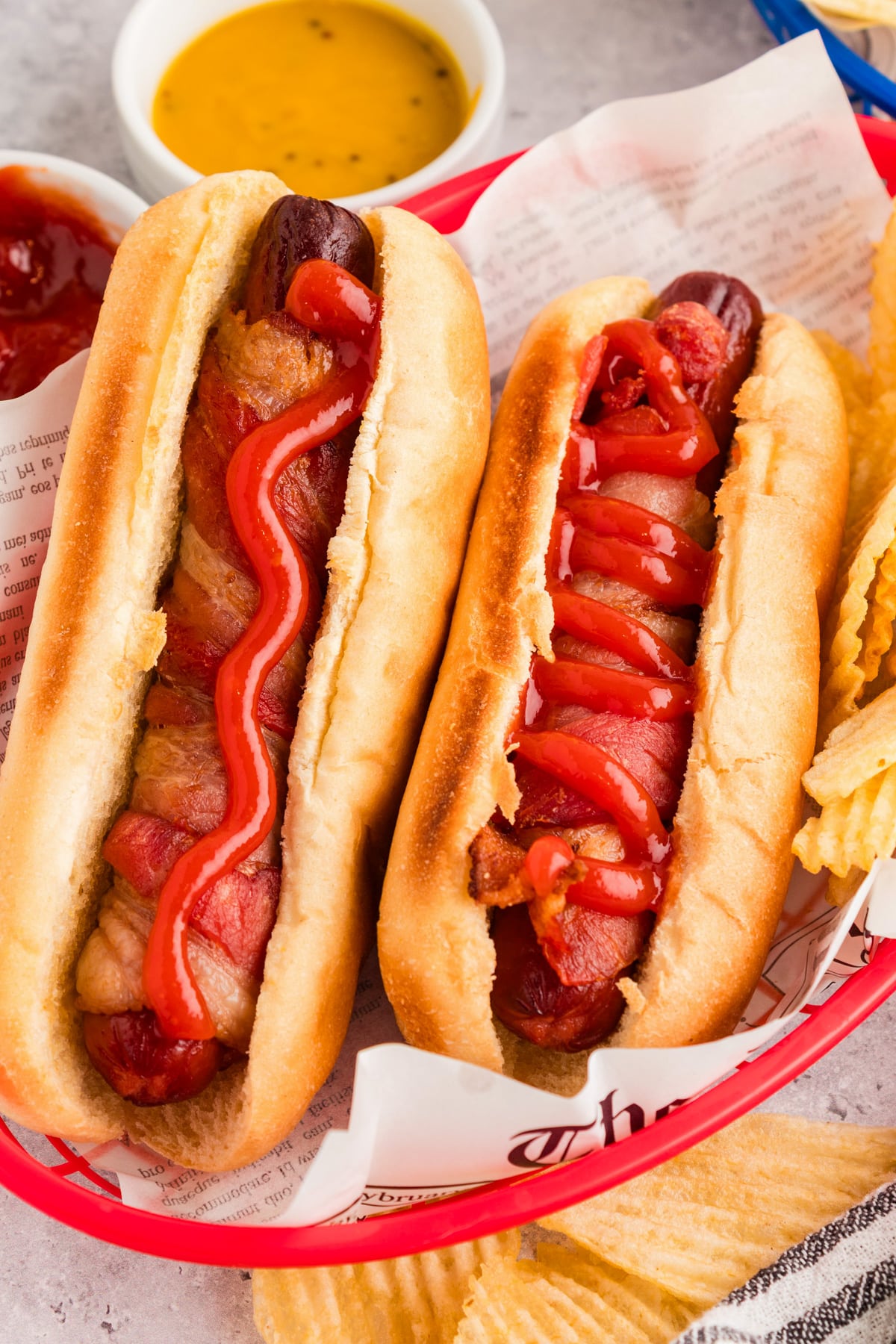 Two bacon wrapped hot dogs in buns, each topped with ketchup, served in a red basket with a side of potato chips and dipping sauces in the background.