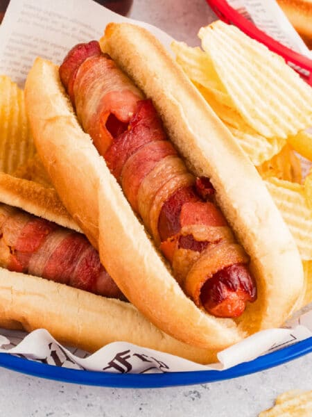Two bacon-wrapped air fryer hot dogs in buns served in a blue basket with wavy potato chips on the side.