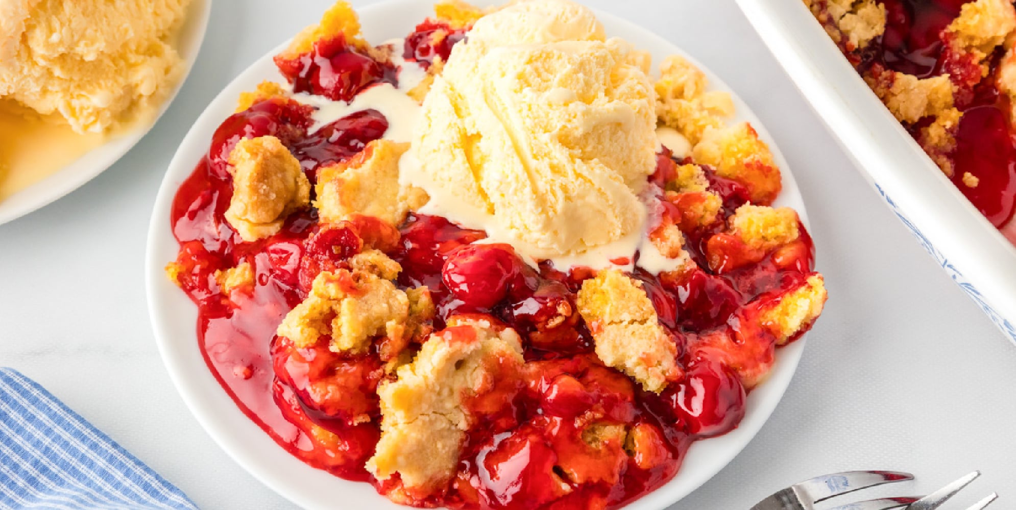 A wide view of a plate of cherry dump cake topped with a scoop of vanilla ice cream on a white surface.