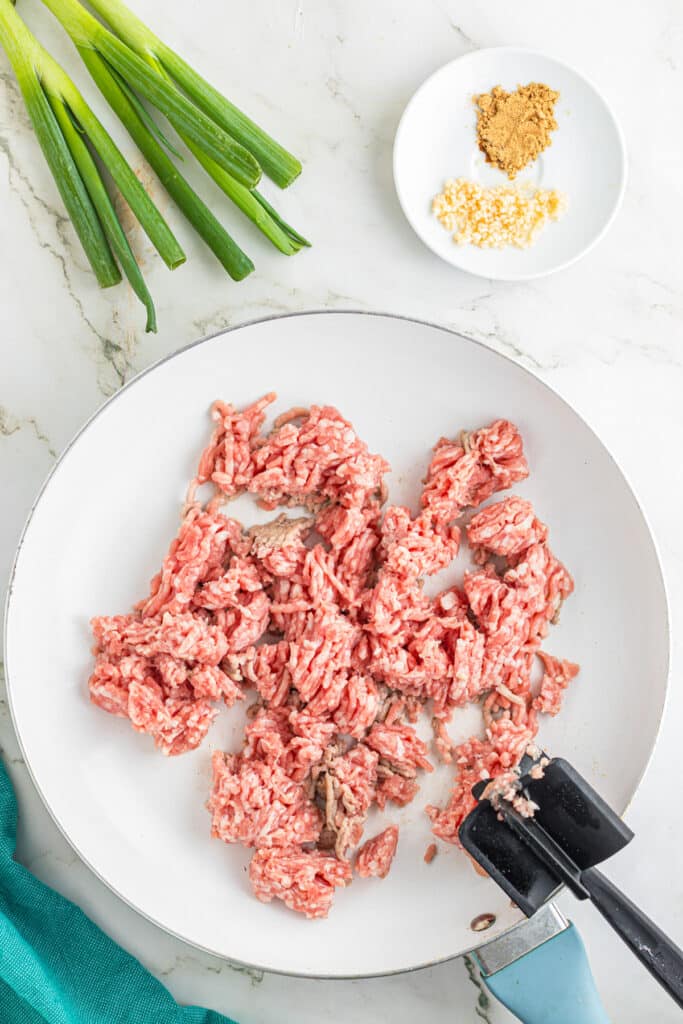 Ground pork being browned in a pan for egg roll in a bowl with spices nearby on the counter.