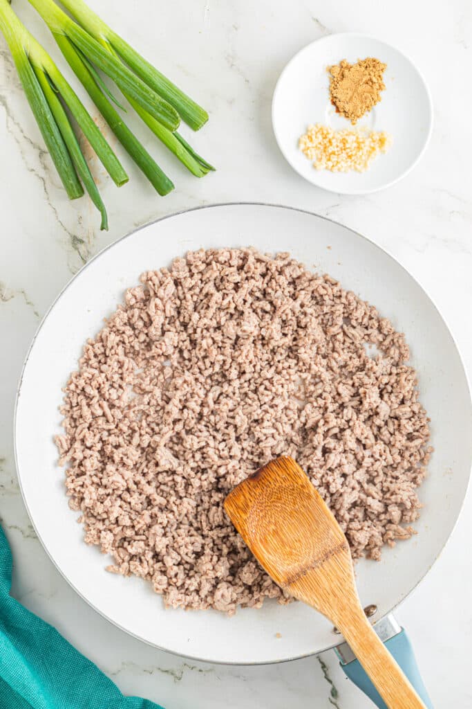 Cooked ground pork in a skillet being moved around the pan with ginger, garlic and green onion nearby to make egg roll in a bowl.