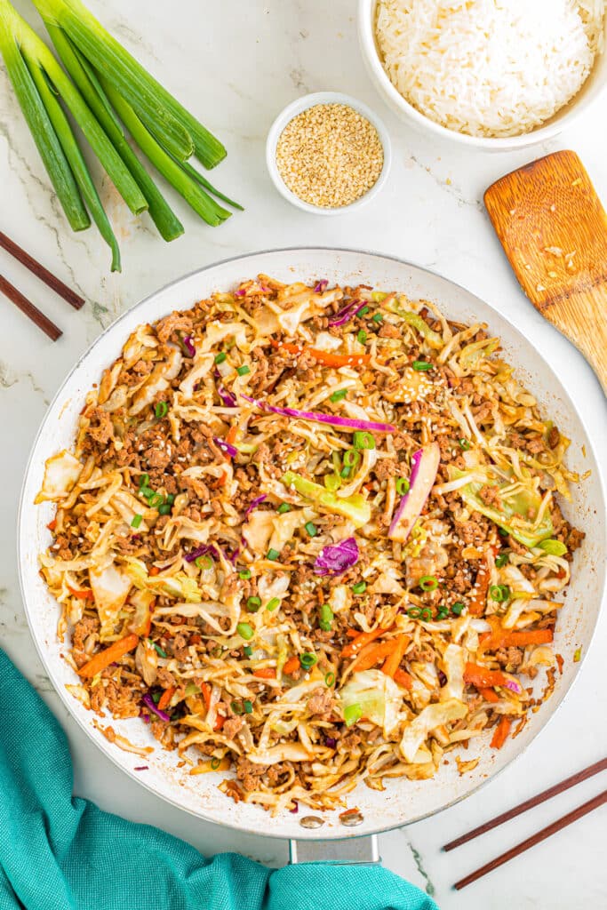 A pan filled with ground pork egg roll in a bowl with a bowl of rice, green onions and sesame seeds on the counter nearby.