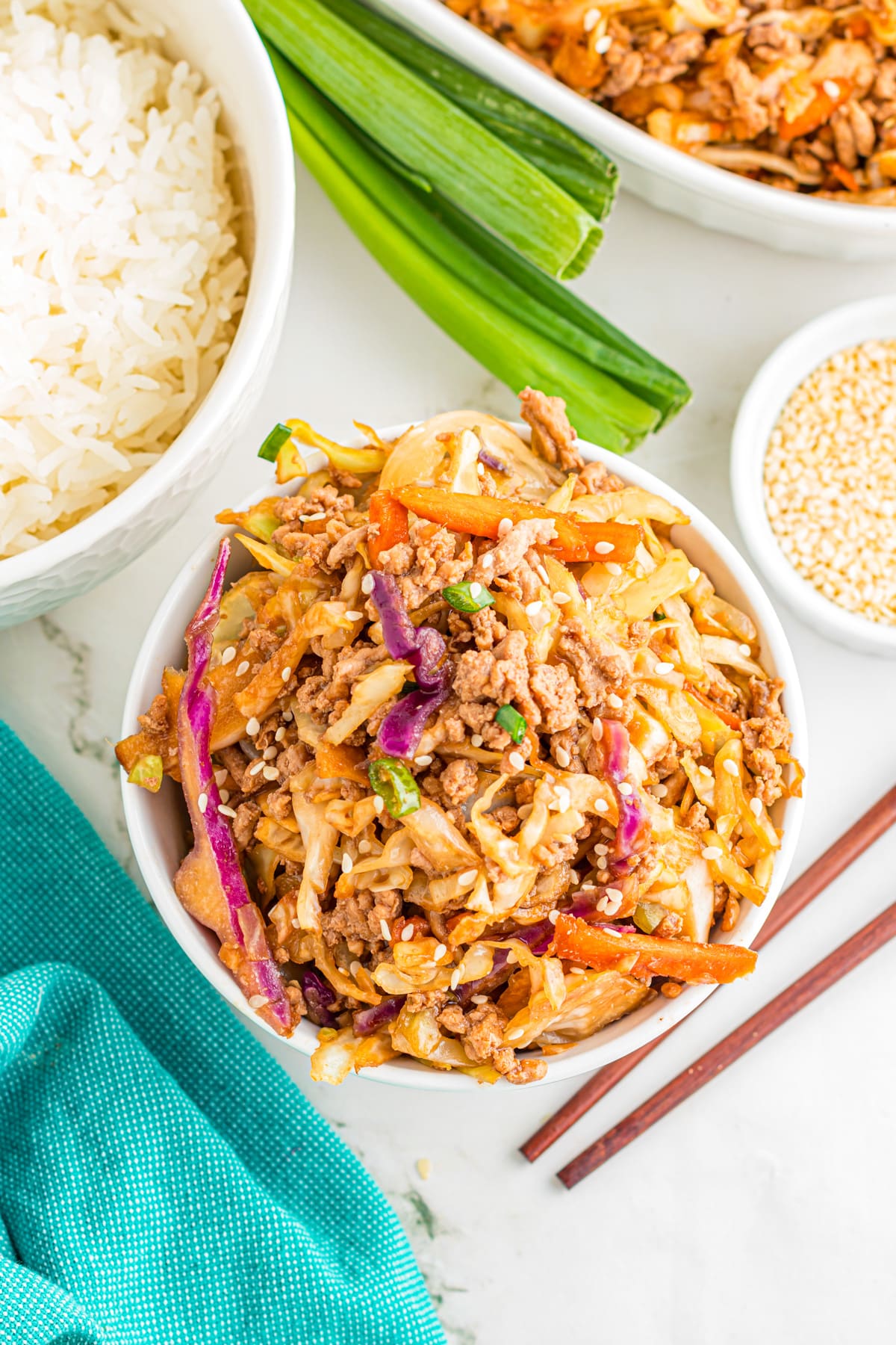 A bowl of ground pork egg roll in a bowl with cabbage, carrots, and green onions, served next to a bowl of white rice, sesame seeds, green onions, and wooden chopsticks.