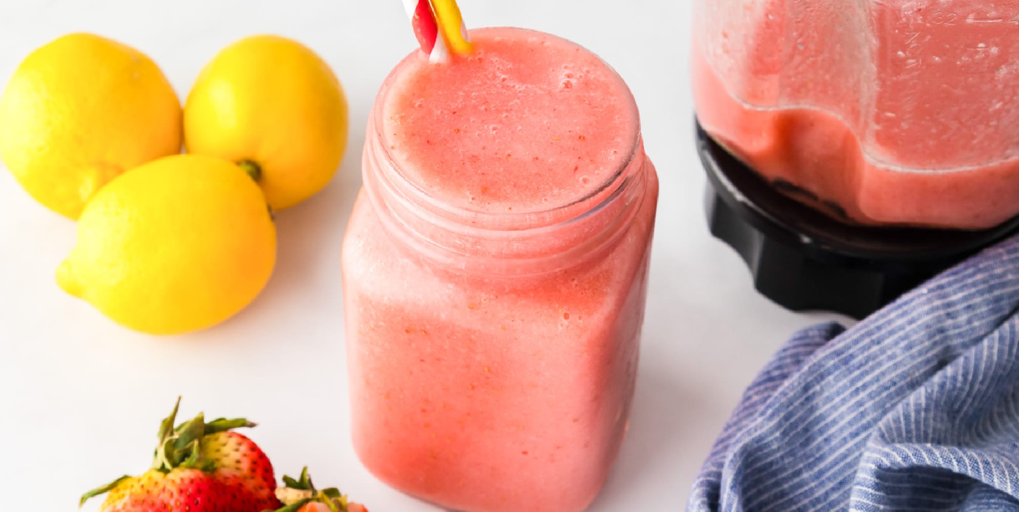 A glass jar filled with pink smoothie and a striped straw next to lemons, strawberries, a blender, and a blue-striped cloth.