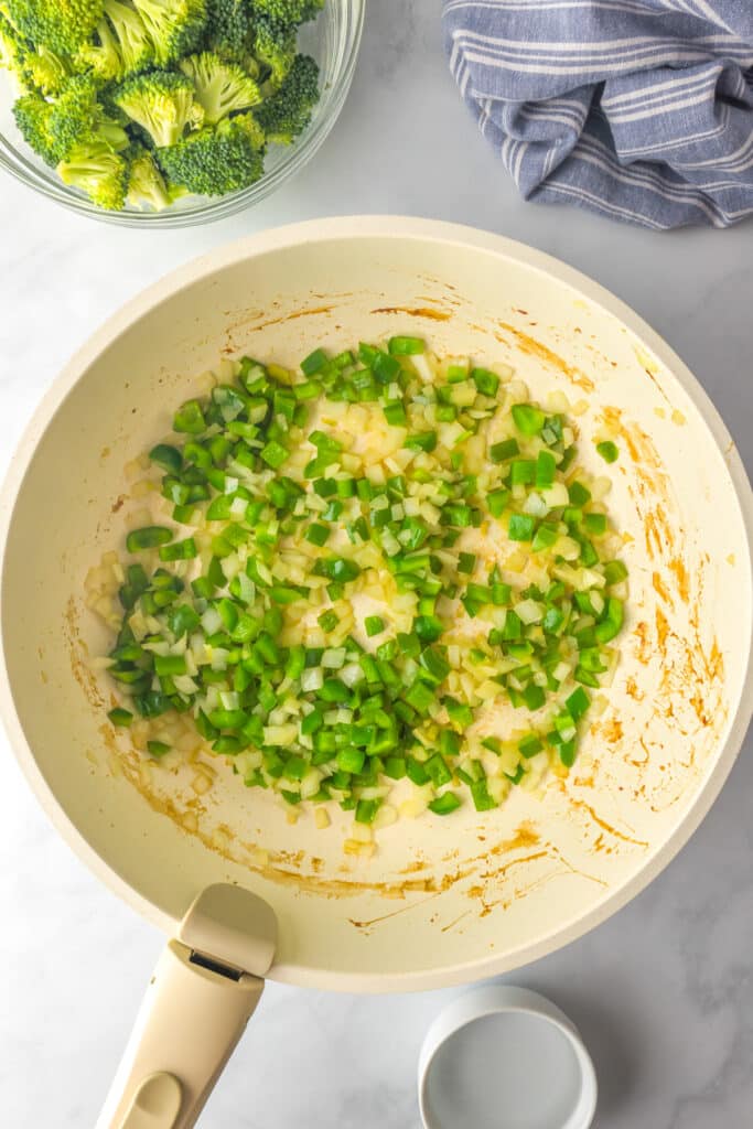 Chopped green bell pepper, onion and garlic cooking in a pan with broccoli in a bowl nearby.