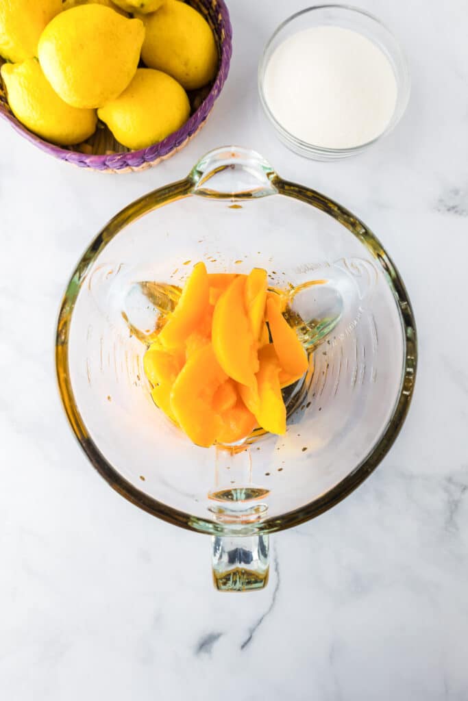A glass blender base with sliced peaches after defrosting with a small bowl of sugar, and a small basket of lemons nearby on the counter.