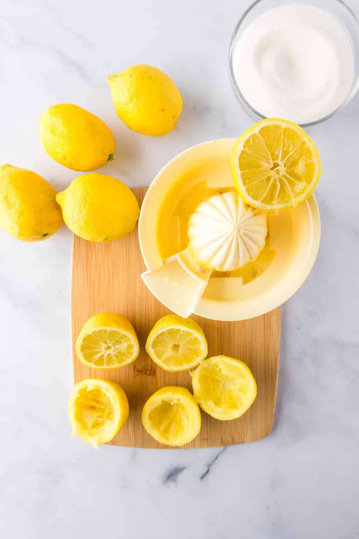 Whole and squeezed lemons on a cutting board next to a manual juice squeezer in the process of being squeezed.