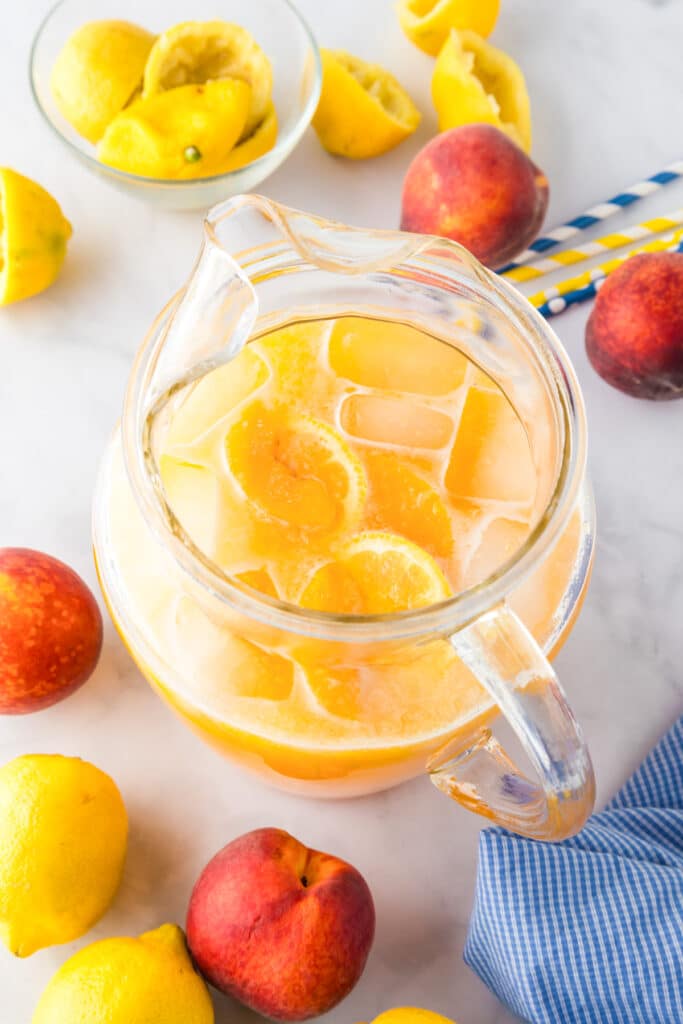 A glass pitcher filled with iced peach lemonade sits on a counter surrounded by more fresh peaches and lemons.
