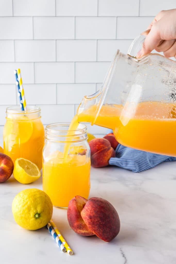 A hand pours peach lemonade from a pitcher into a glass with another full cup in the background.
