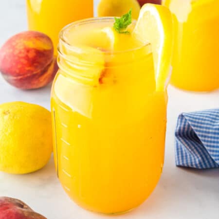 A mason jar filled with peach lemonade with fresh peaches and lemons on the counter nearby.