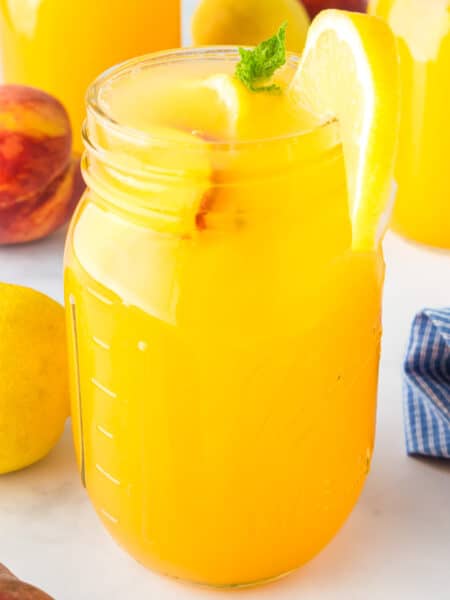 A mason jar filled with peach lemonade with fresh peaches and lemons on the counter nearby.