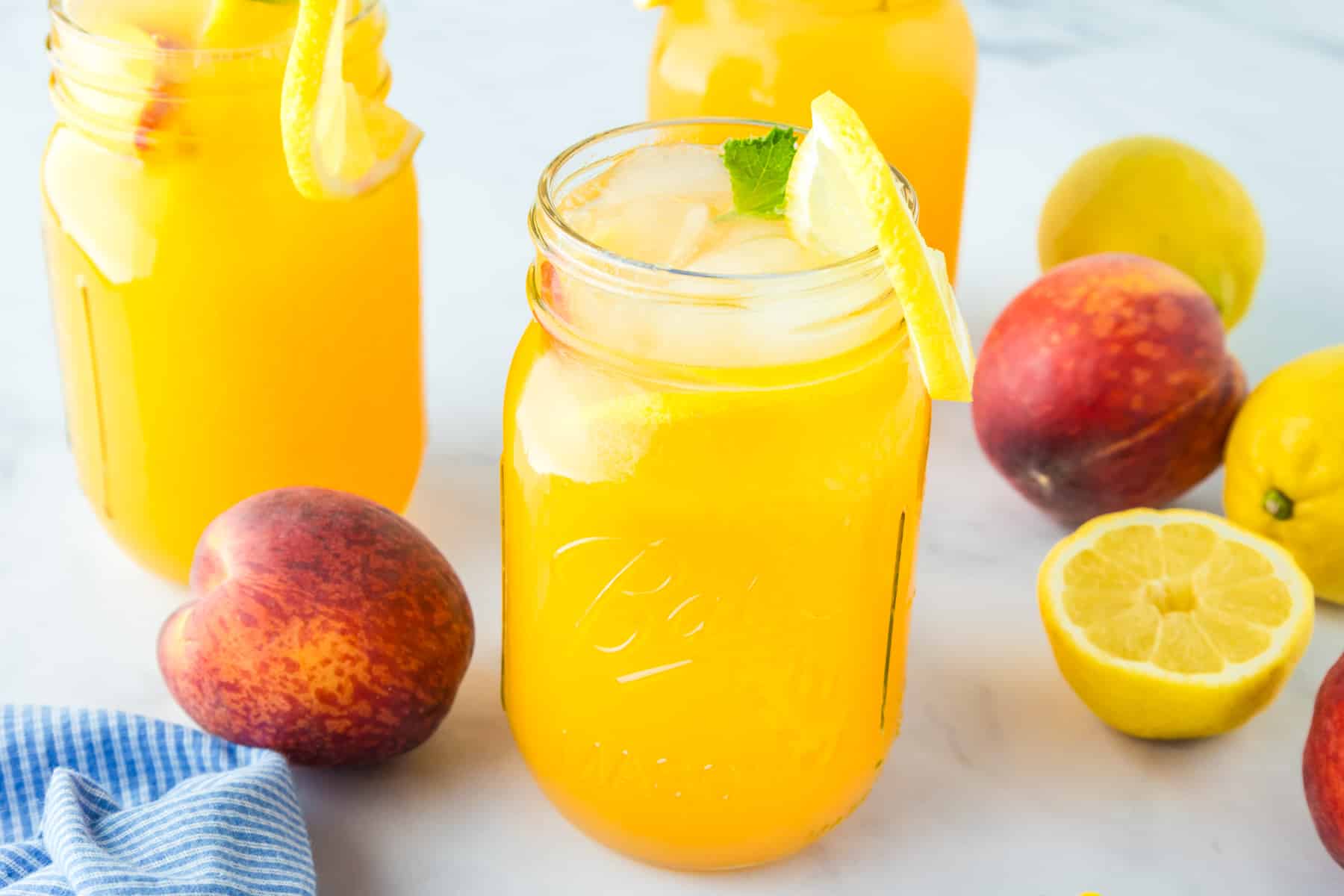 Three mason jars filled with peach-colored iced drinks garnished with lemon slices, surrounded by fresh peaches and lemons on a white surface.