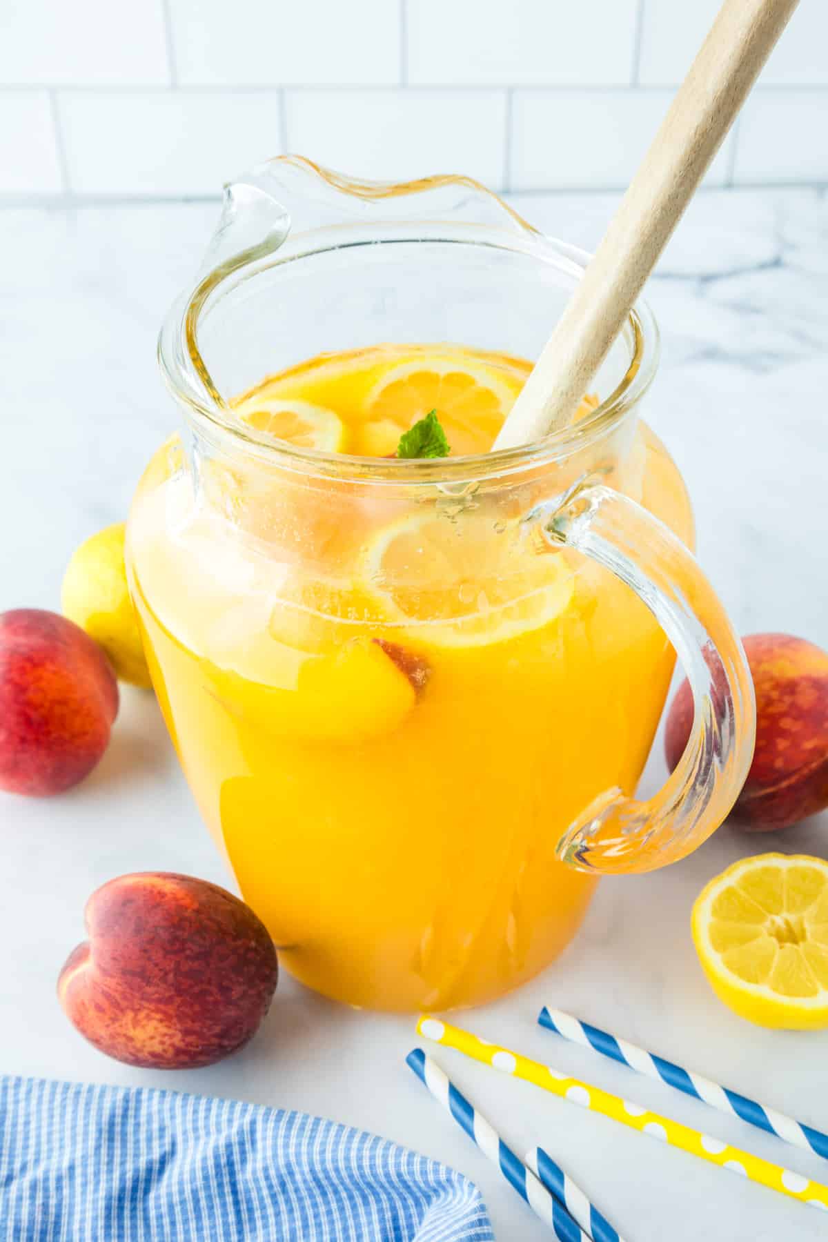 A glass pitcher filled with peach lemonade, garnished with lemon slices and mint, sits on a marble surface with fresh peaches, a lemon, and straws nearby.