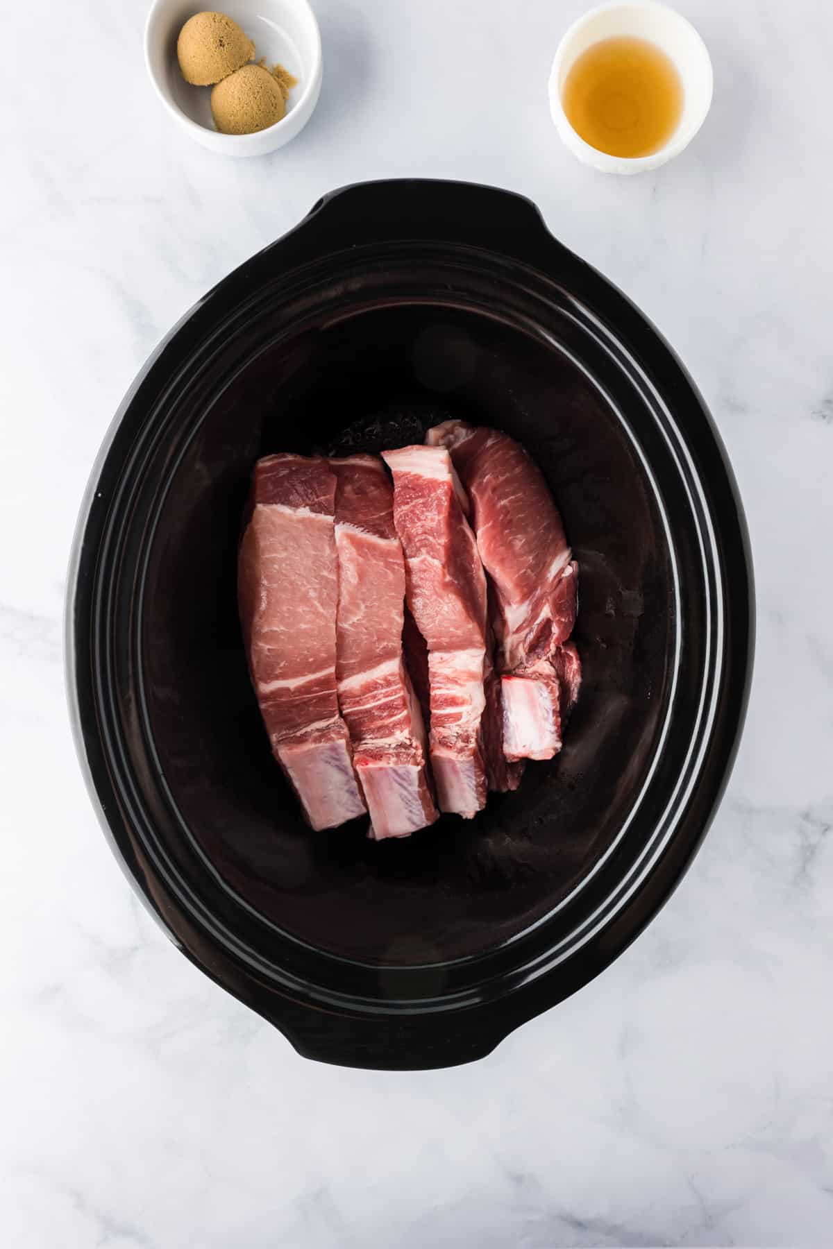 Raw country style loin pork ribs arranged in a black slow cooker, with small bowls of brown sugar and apple cider vinegar on a white marble surface nearby.