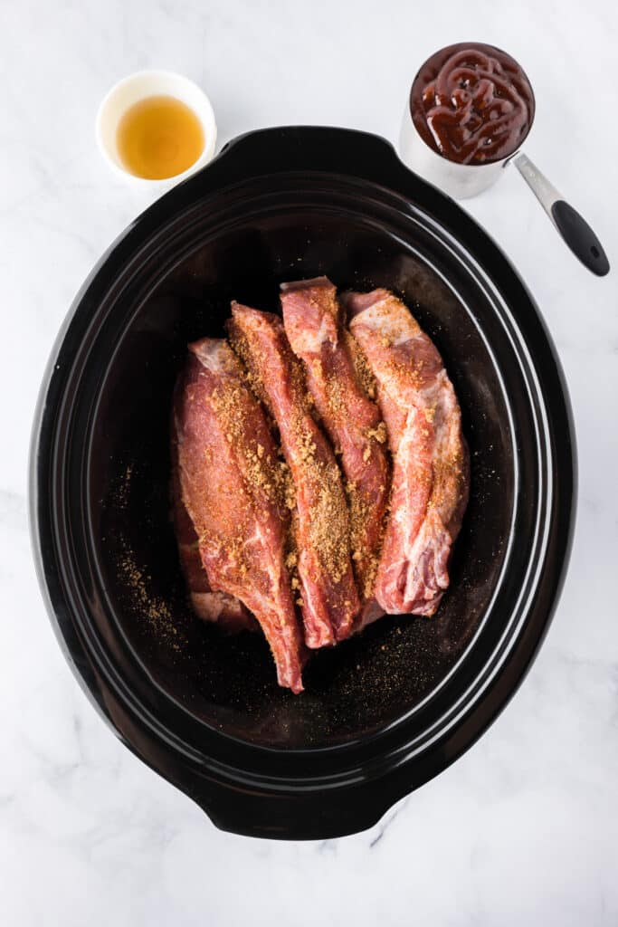 Raw pork loin country style ribs coated with dry rub seasoning after flipping the ribs in a slow cooker with bbq sauce and apple cider vinegar nearby on the counter.