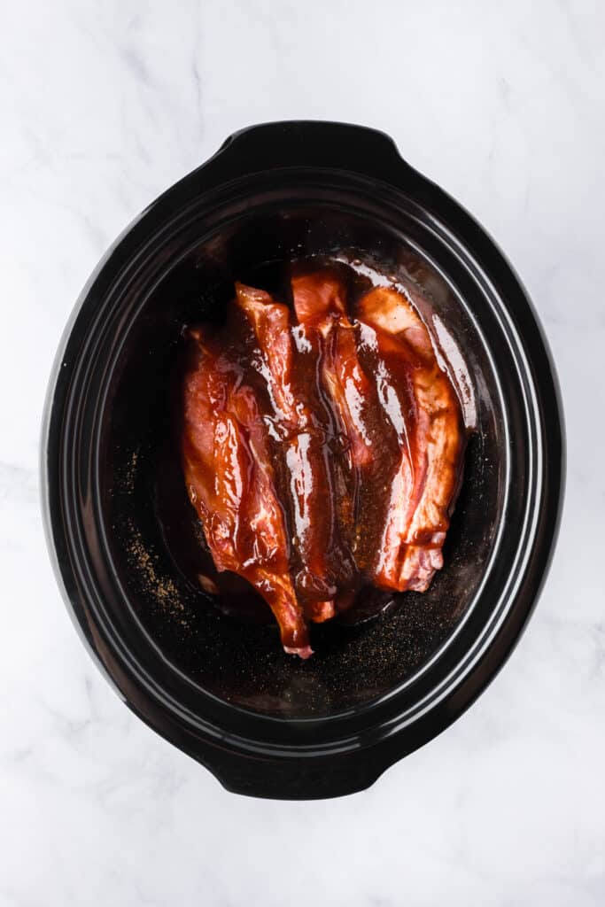 Raw loin country style pork ribs coated in barbecue sauce placed in a slow cooker.