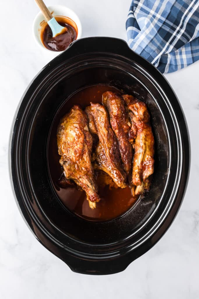 Cooked loin country style pork ribs coated in barbecue sauce in a slow cooker with more bbq sauce in a bowl nearby.