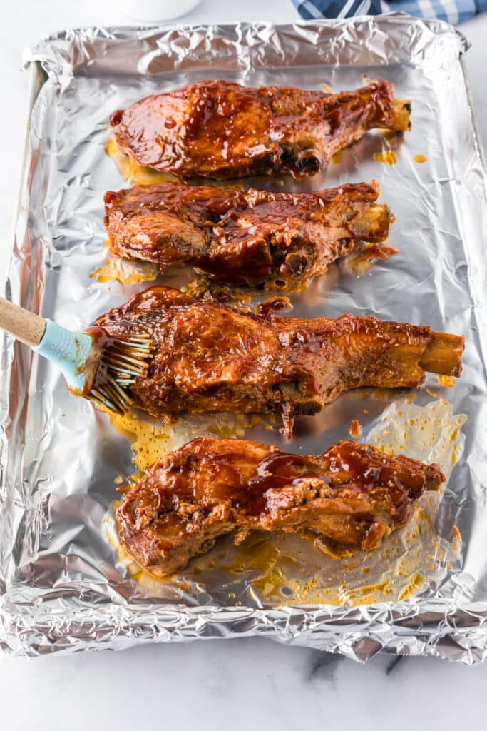 Four cooked loin country style pork ribs on a foil-lined baking sheet being brushed with barbecue sauce.
