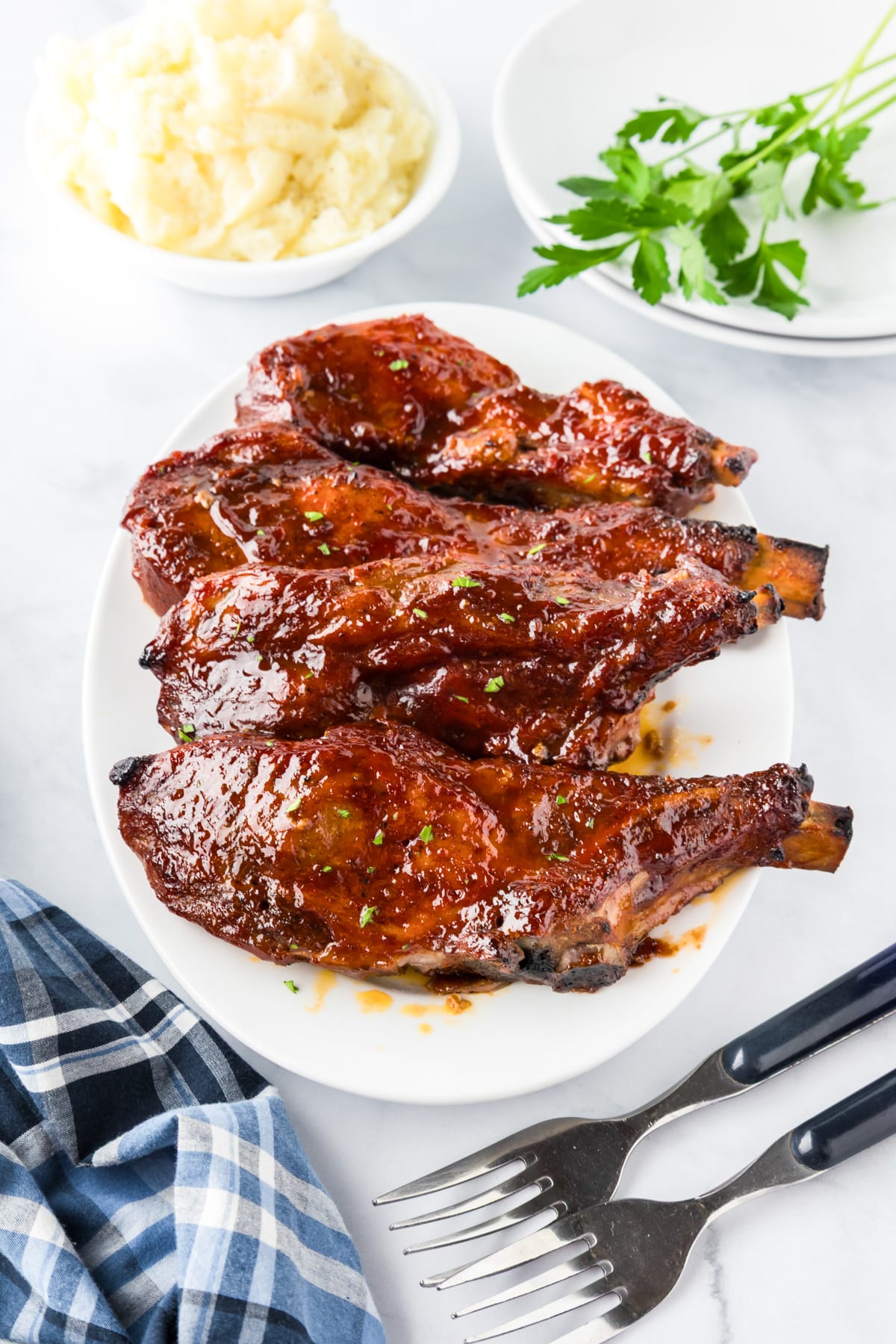 Four glazed barbecue loin country style ribs on a plate with a bowl of mashed potatoes, parsley, and two forks nearby.
