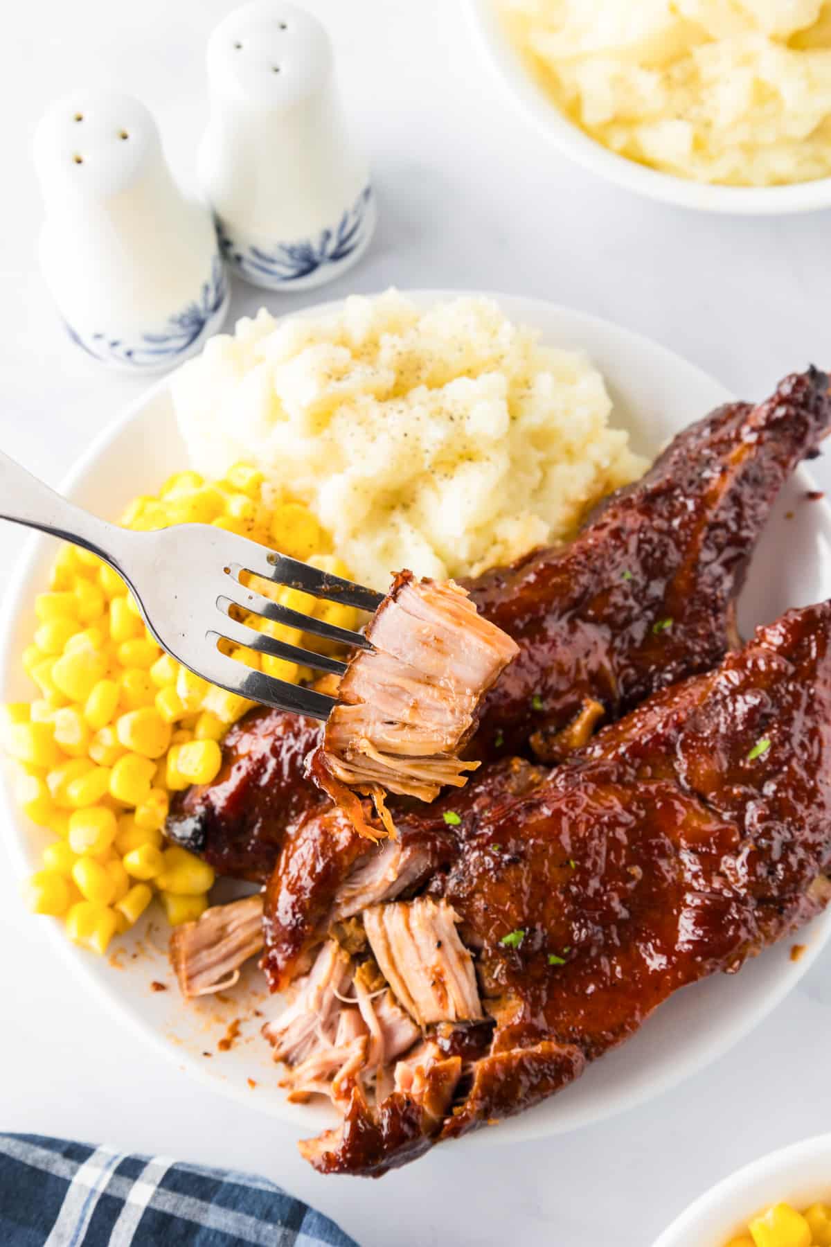 Plate with barbecued slow cooker loin country style pork ribs, mashed potatoes with pepper, and corn; a fork holds a piece of rib meat.