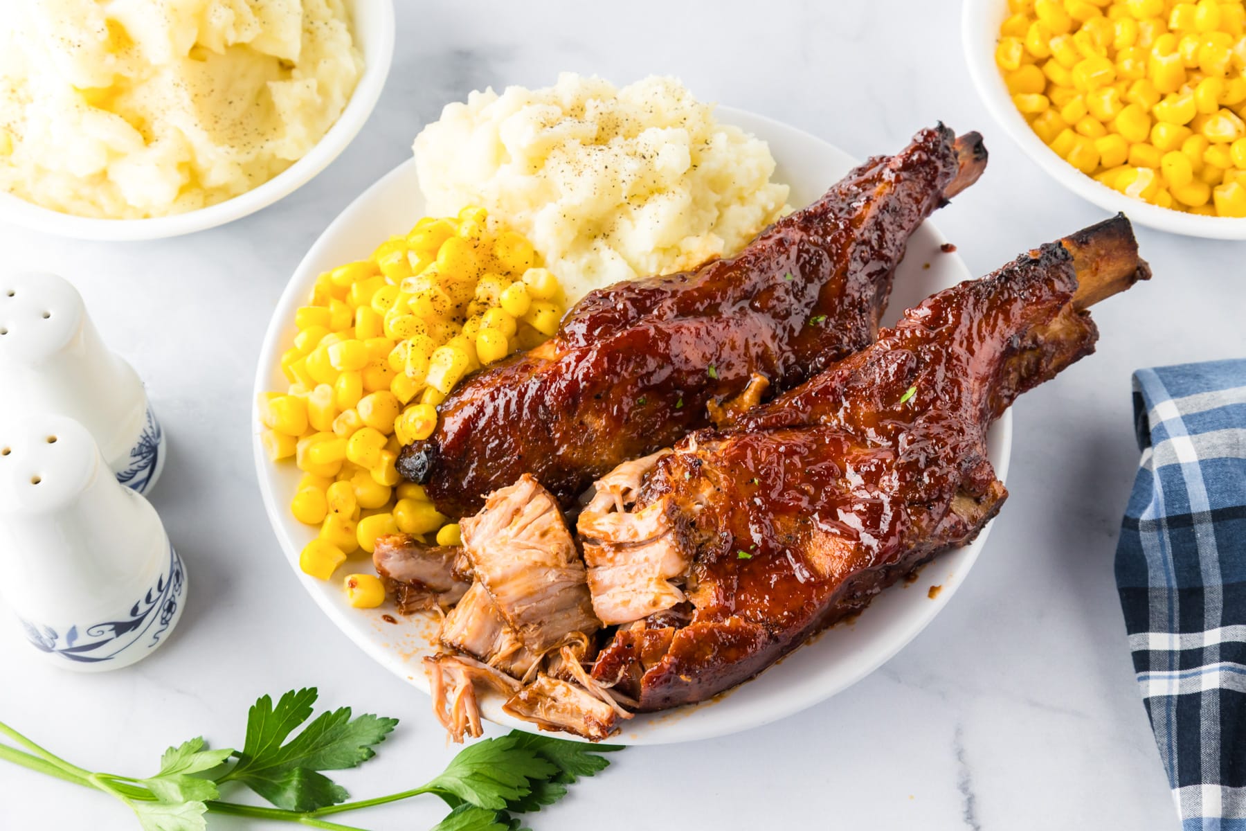 A plate with two barbecued loin country styke pork ribs, mashed potatoes with pepper, and corn on a dinner table with the inside of the juicy ribs showing.