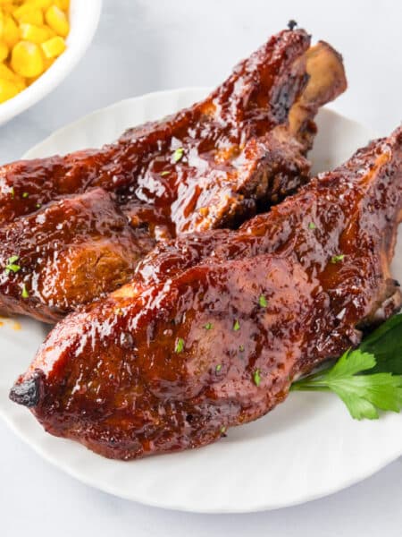 Two glazed slow cooker loin country style pork ribs on a plate cooked in bbq sauce with a bowl of corn in the background.