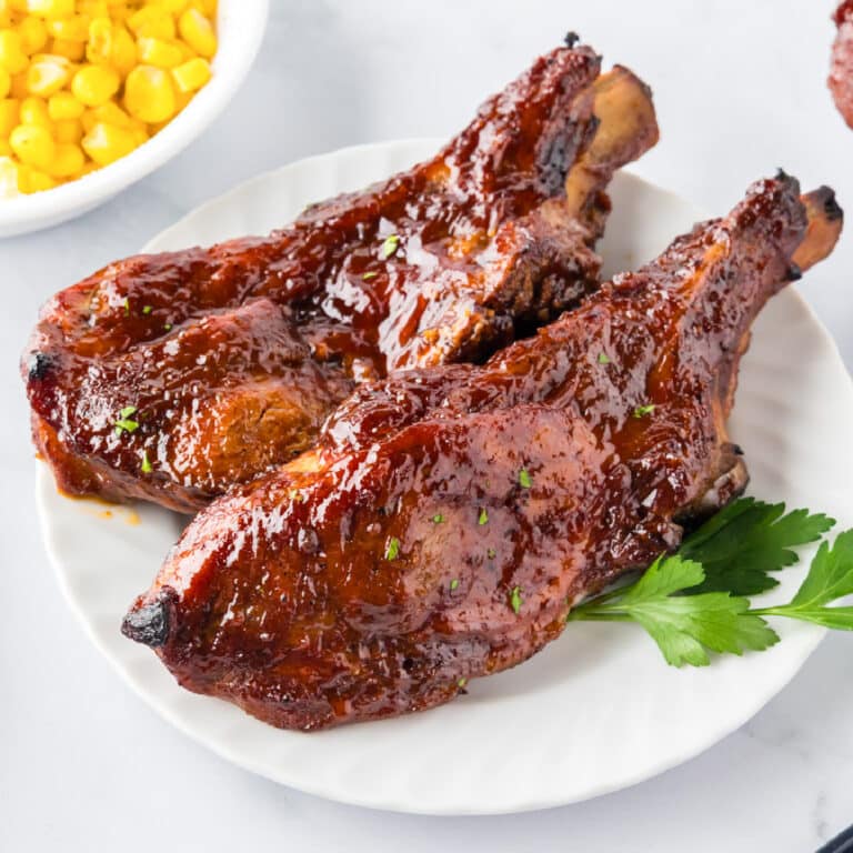 Two glazed slow cooker loin country style pork ribs on a plate cooked in bbq sauce with a bowl of corn in the background.
