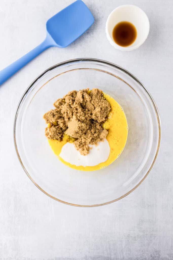 A glass bowl with wet ingredients including beaten eggs, brown sugar, and white sugar, next to a blue spatula and a small bowl of vanilla extract for zucchini brownies.