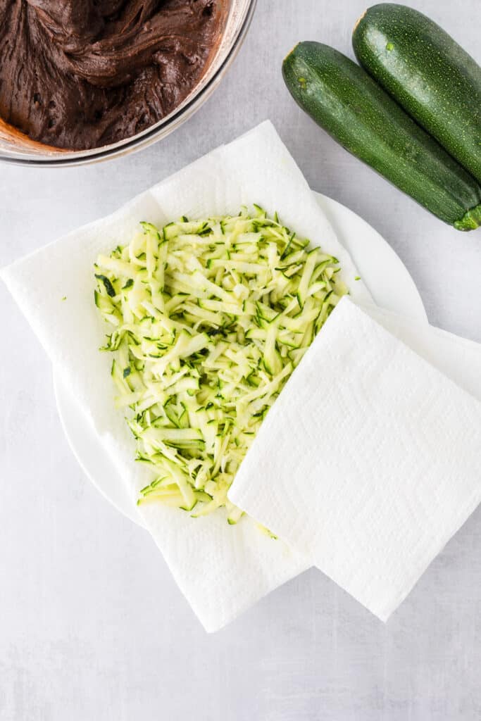 Shredded zucchini on a paper towel-lined plate being patted dry for zucchini brownies next to two whole zucchinis and a bowl of chocolate brownie batter.