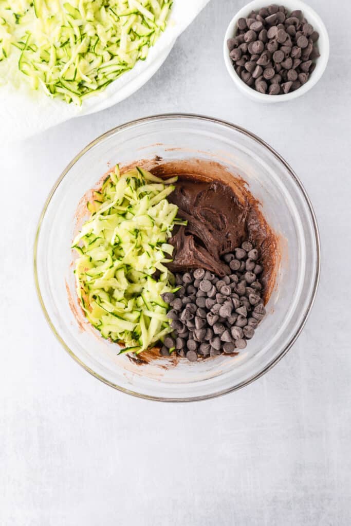 A glass bowl with chocolate zucchini brownie batter, shredded zucchini , and chocolate chips being mixed together.