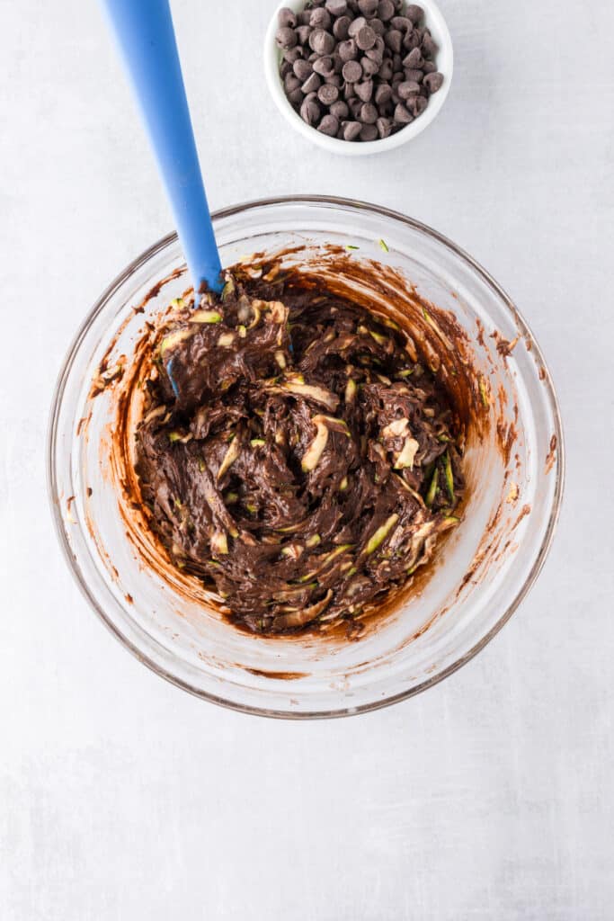 A glass bowl with chocolate zucchini brownie batter, shredded zucchini , and chocolate chips after mixing.