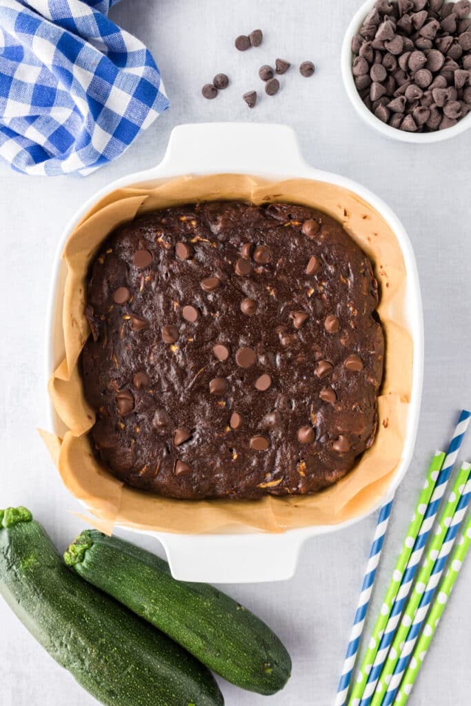 A square baking dish lined with parchment paper holds unbaked chocolate zucchini batter topped with chocolate chips for zucchini brownies.