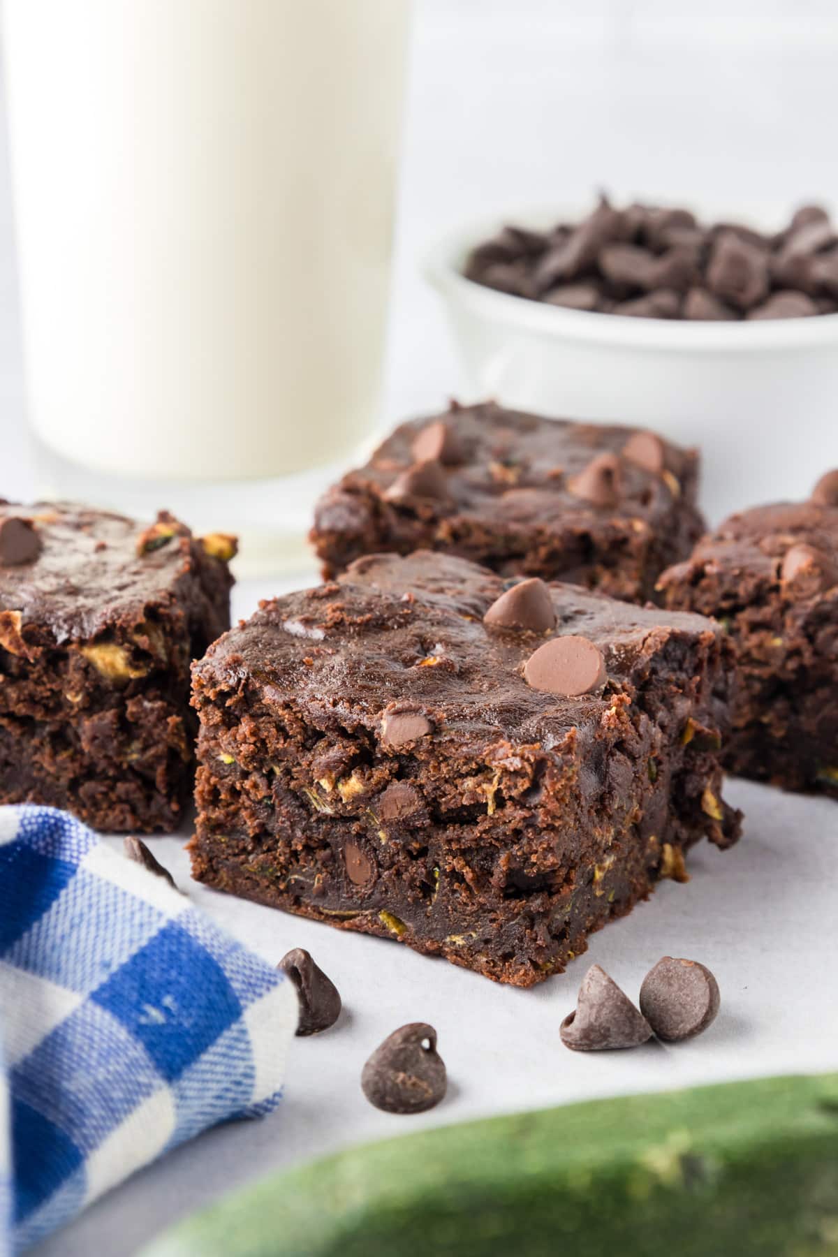 Three chocolate chip brownies are arranged on a piece of parchment paper by focused on the center brownie with a glass of milk, zucchini and a bowl of chocolate chips nearby on the table.