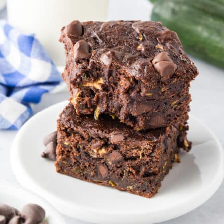 Two chocolate chip zucchini brownies are stacked on a white plate, with the top brownie missing a bite.