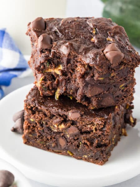 Two chocolate chip zucchini brownies are stacked on a white plate, with the top brownie missing a bite.
