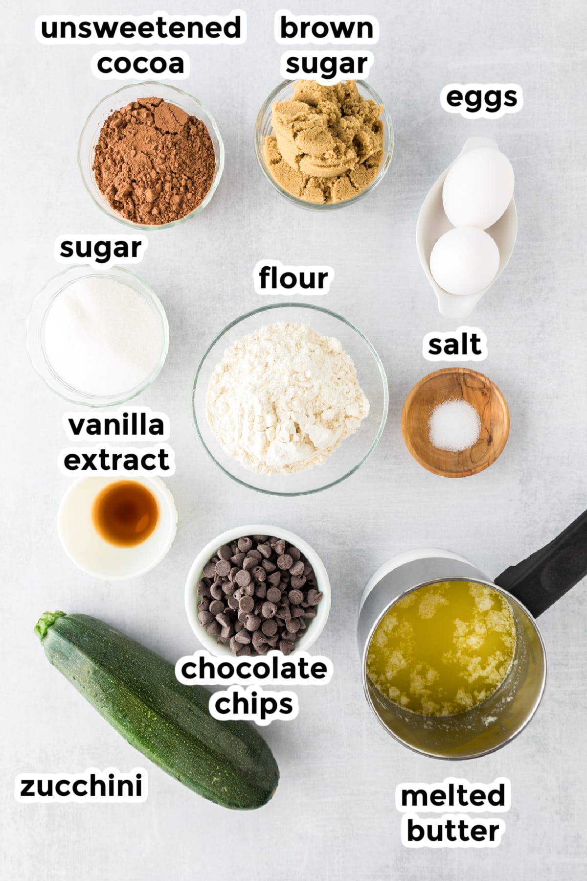 Ingredients for zucchini brownies in bowls with text titles on a counter from overhead.