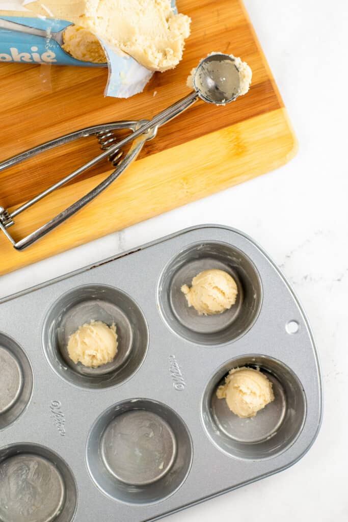 Portions of cookie dough being placed in a greased muffin tin for apple cookie cups, with a scoop and cookie dough packaging on a cutting board nearby.