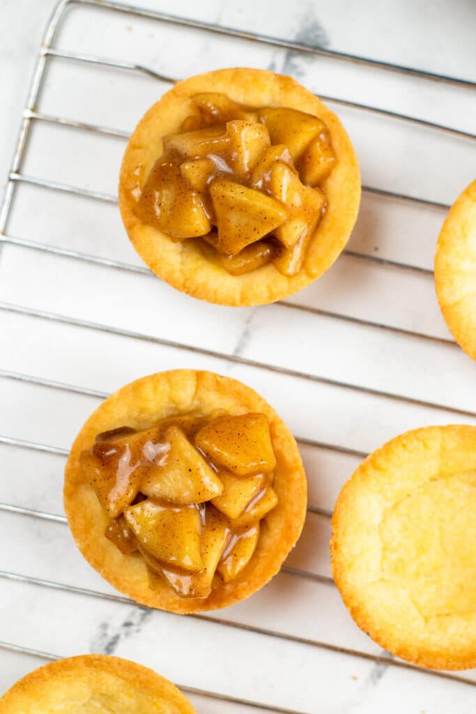 Two mini filled apple cookie cups on a wire rack next to more cookie shells.