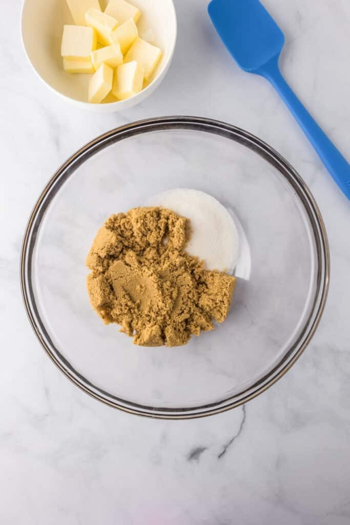 A glass mixing bowl with brown and white sugar being mixed with a small bowl of cubed butter on the counter nearby for blondies.