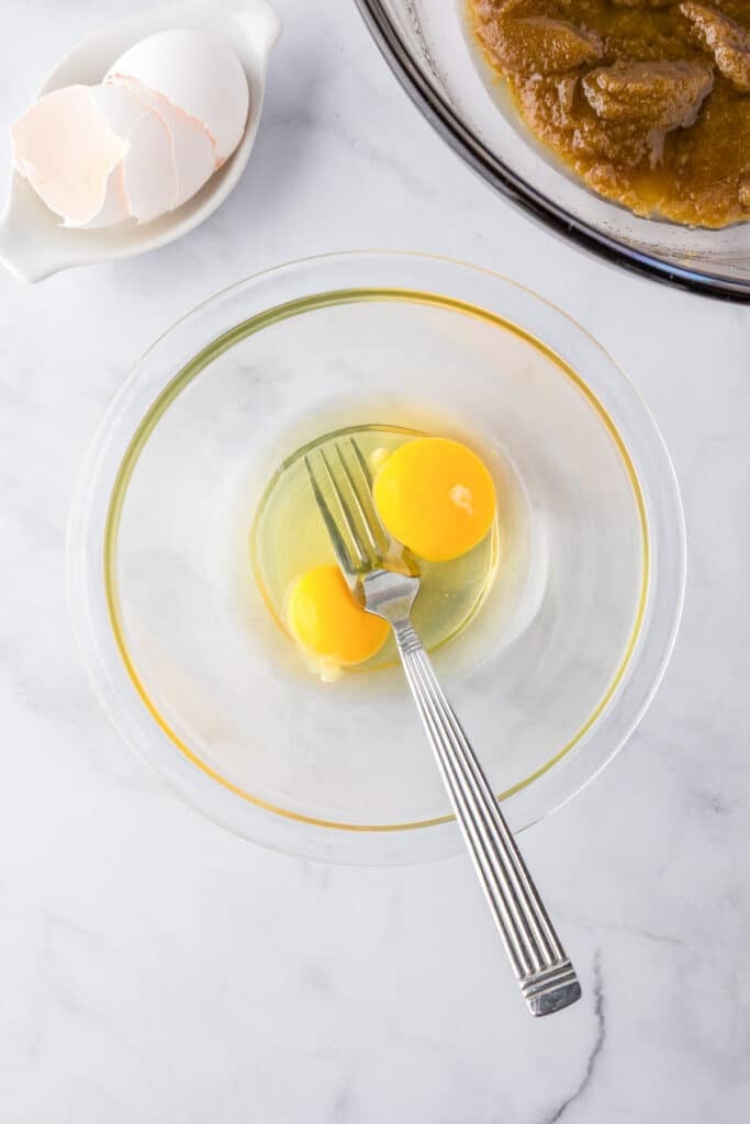 A clear bowl with two raw eggs and a metal fork for mixing them for blondies.