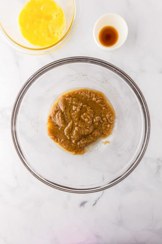 A glass bowl with a brown sugar and white sugar being dissolved into melted butter with vanilla extract and beaten eggs in bowls nearby for making blondies.