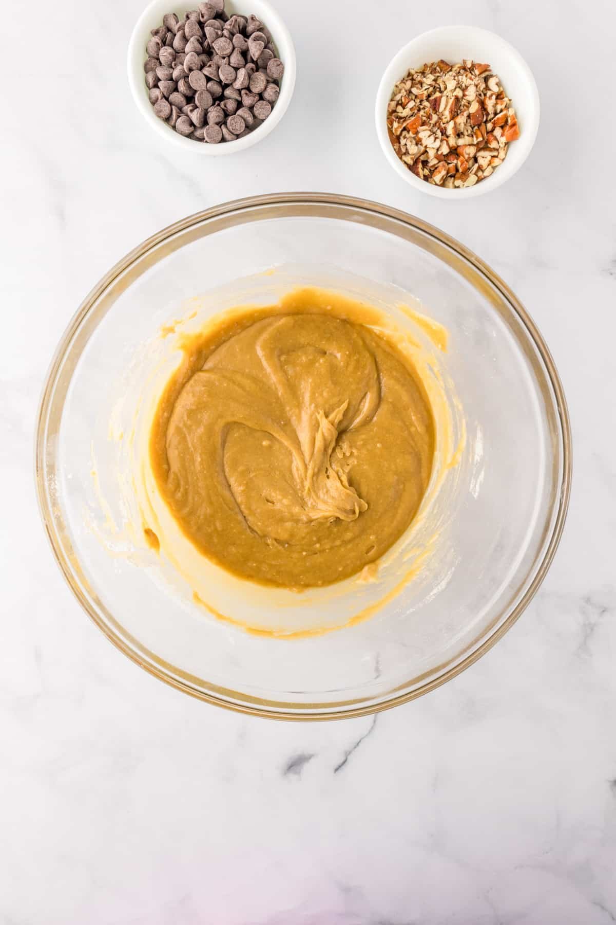 A glass bowl with blondie batter after mixing with chocolate chips and chopped pecans in bowls nearby.