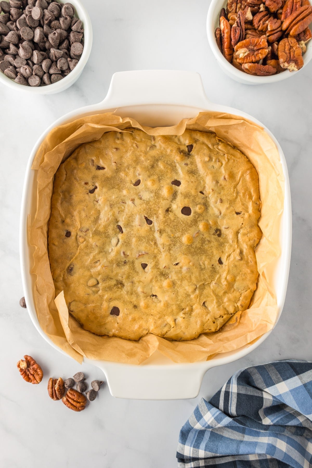 Blondies baked in a square pan with chocolate chips and pecans in bowls on the counter nearby.