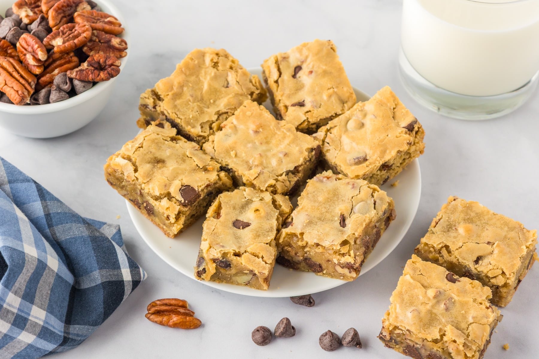 Wide view of a platter of chocolate chip pecan blondies on a counter next to a glass of milk, and a bowl of nuts and chocolate chips.