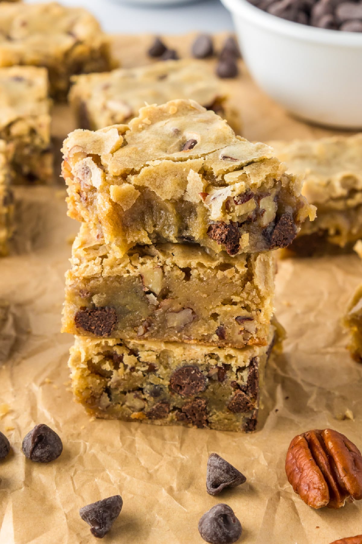 Three blondie bars with chocolate chips and pecans are stacked on parchment paper, the top bar missing a bite.