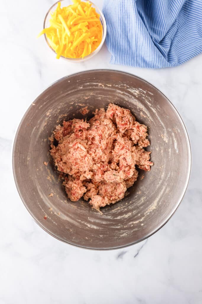 A metal mixing bowl filled with raw ground meat mixture sits on a marble surface next to a bowl of shredded cheddar cheese and a blue cloth.