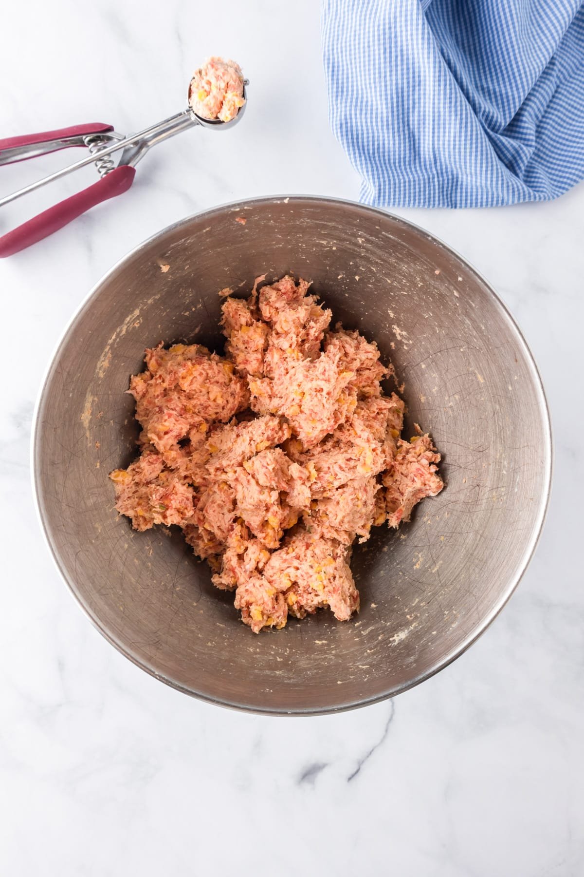 A metal mixing bowl filled with raw sausage ball mixture with a cookie scoop full of one ball nearby.