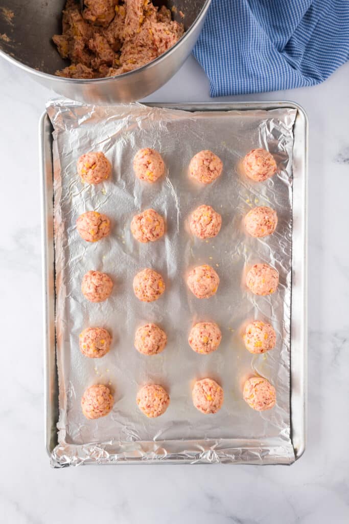 A baking sheet lined with foil holds 20 evenly spaced raw cheddar bay sausage balls.